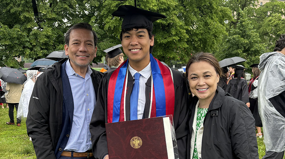 Matthew Raroque wearing his undergraduate graduation cap and gown outside with his parents